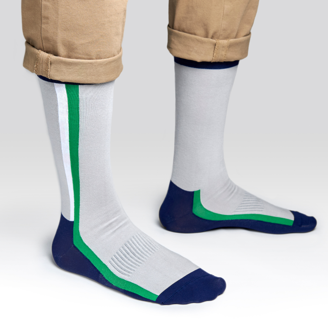 A person wearing light gray socks with navy blue toes and heels, a white vertical stripe, and a green accent stripe. The person’s khaki pants are rolled up above the ankles. The background is plain and light gray.