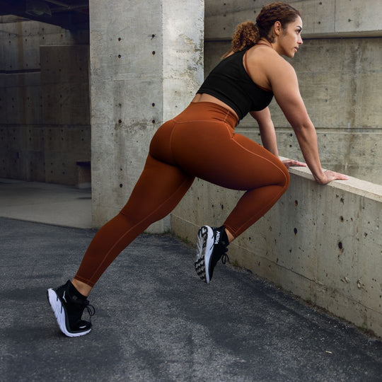 A woman in a black crop top and rust-colored leggings stretches her legs against a concrete wall outdoors, preparing for exercise. She wears black running shoes and has curly hair tied back.