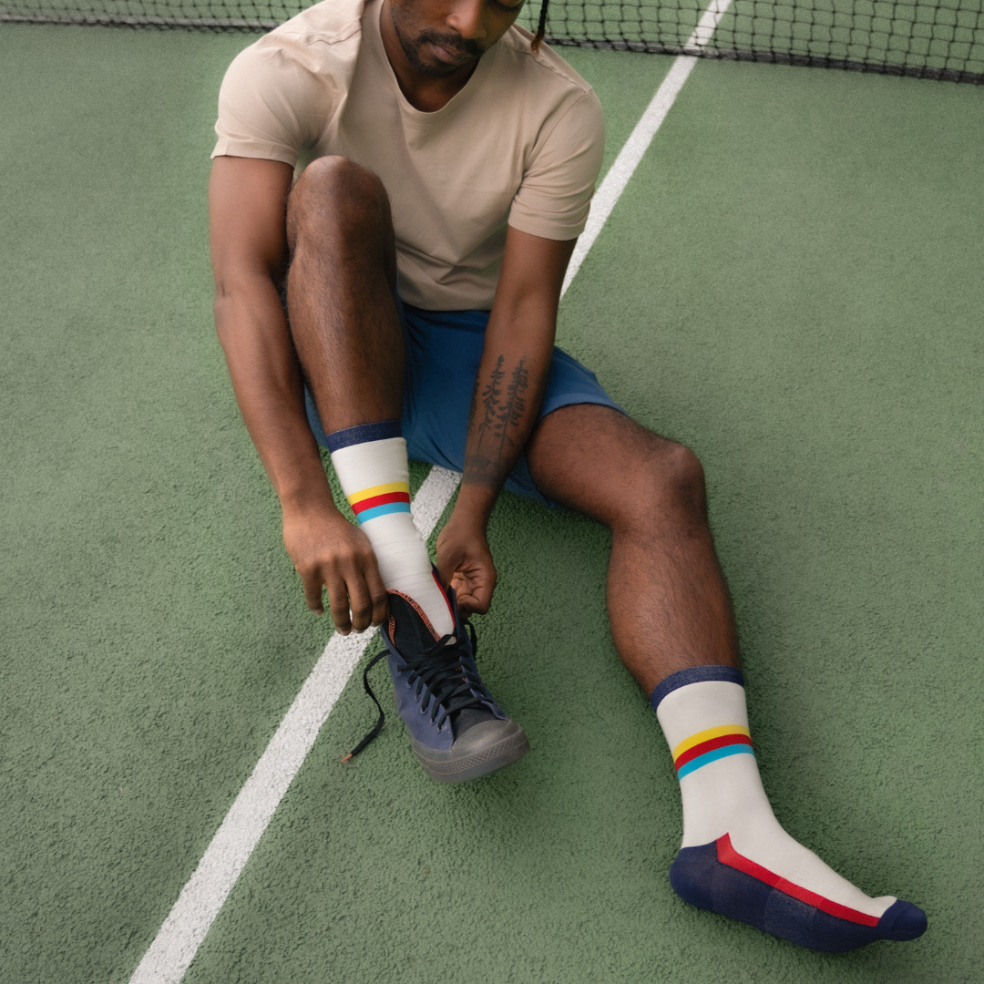 A person sitting on a tennis court ties the laces of a navy sneaker. They wear a beige t-shirt, blue shorts, and white socks with colorful rainbow stripes near the top.