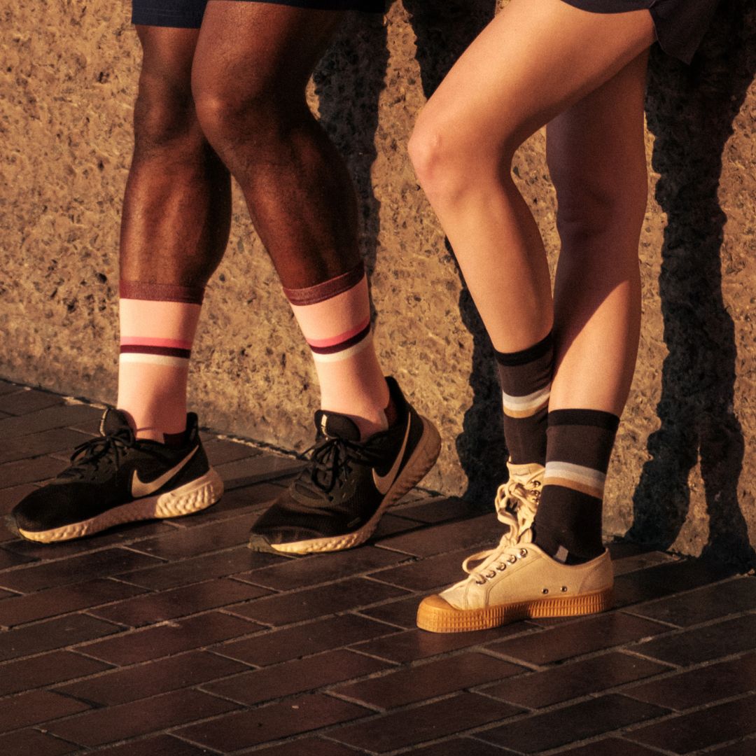 Two people standing side by side against a wall, showing only their legs. Both are wearing shorts, sneakers, and colorful striped socks in shades of pink, black, and tan, with sunlight casting shadows on them.