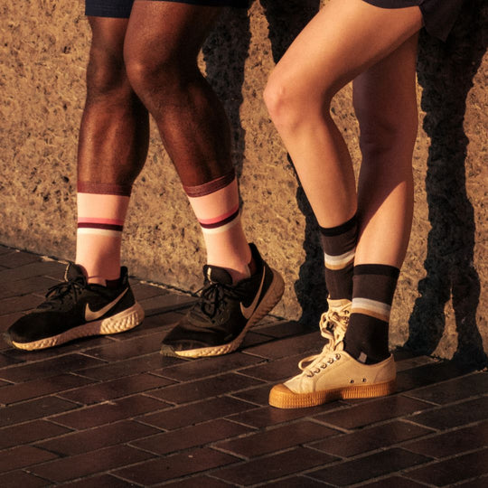 Two people standing side by side against a wall, showing only their legs. Both are wearing shorts, sneakers, and colorful striped socks in shades of pink, black, and tan, with sunlight casting shadows on them.