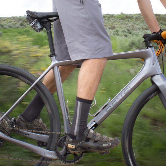 A close-up side view of a person riding a gray Revel bicycle outdoors, wearing gray shorts, black and gray socks, and black cycling shoes, with greenery blurred in the background.