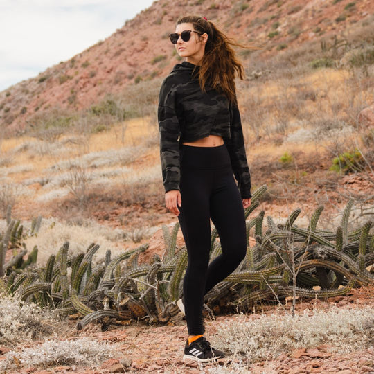 A woman wearing sunglasses, a black camo hoodie, and black leggings stands on a rocky desert trail with cacti around her and hills in the background.