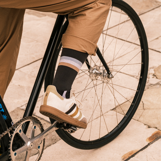 A person wearing tan pants, striped socks, and white sneakers with gold stripes is riding a black bicycle on a tiled pavement. Only their lower leg and part of the bike are visible.