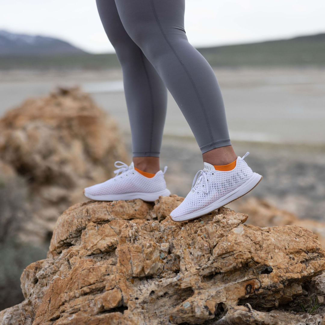 A person wearing gray leggings and white athletic shoes with orange socks stands on a rocky surface outdoors, with a blurred natural landscape in the background.