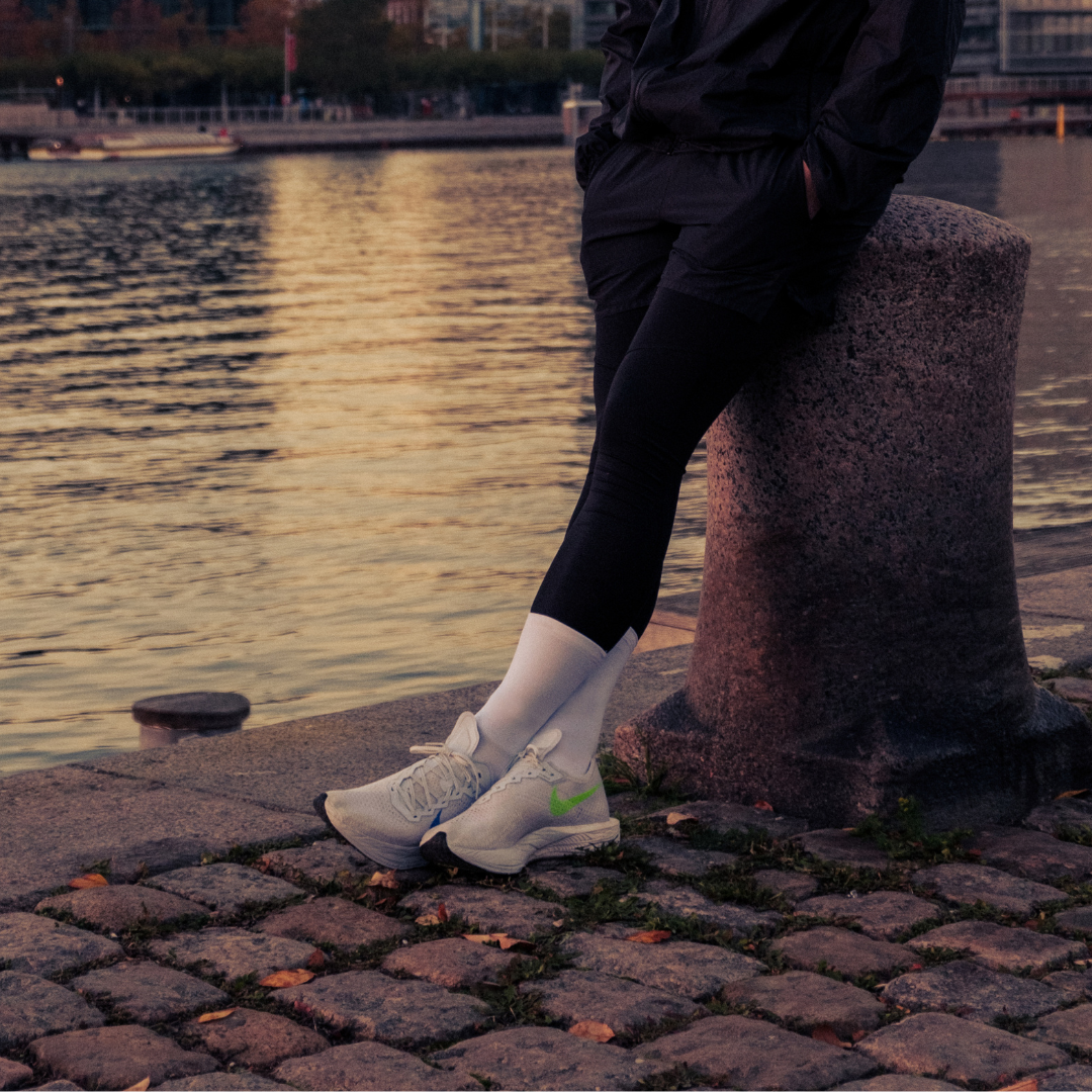 A person in black athletic wear and white sneakers leans against a stone post on a cobblestone path by the waterfront at sunset. Only their lower body is visible.