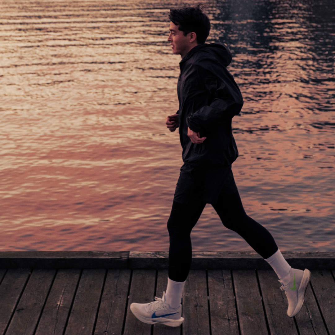 A person jogs along a wooden boardwalk beside calm water at sunset, wearing dark athletic clothes, white sneakers, and white socks.