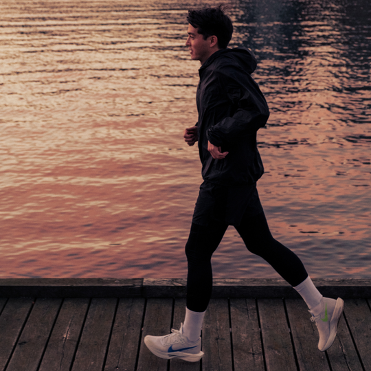 A person jogs along a wooden boardwalk beside calm water at sunset, wearing dark athletic clothes, white sneakers, and white socks.
