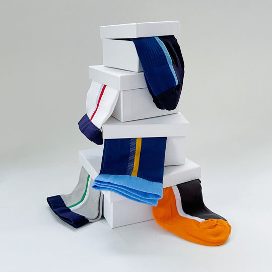 A stack of white boxes with various colorful socks draped over them, including blue, orange, black, gray, and white socks with different patterns and stripes, set against a plain light background.
