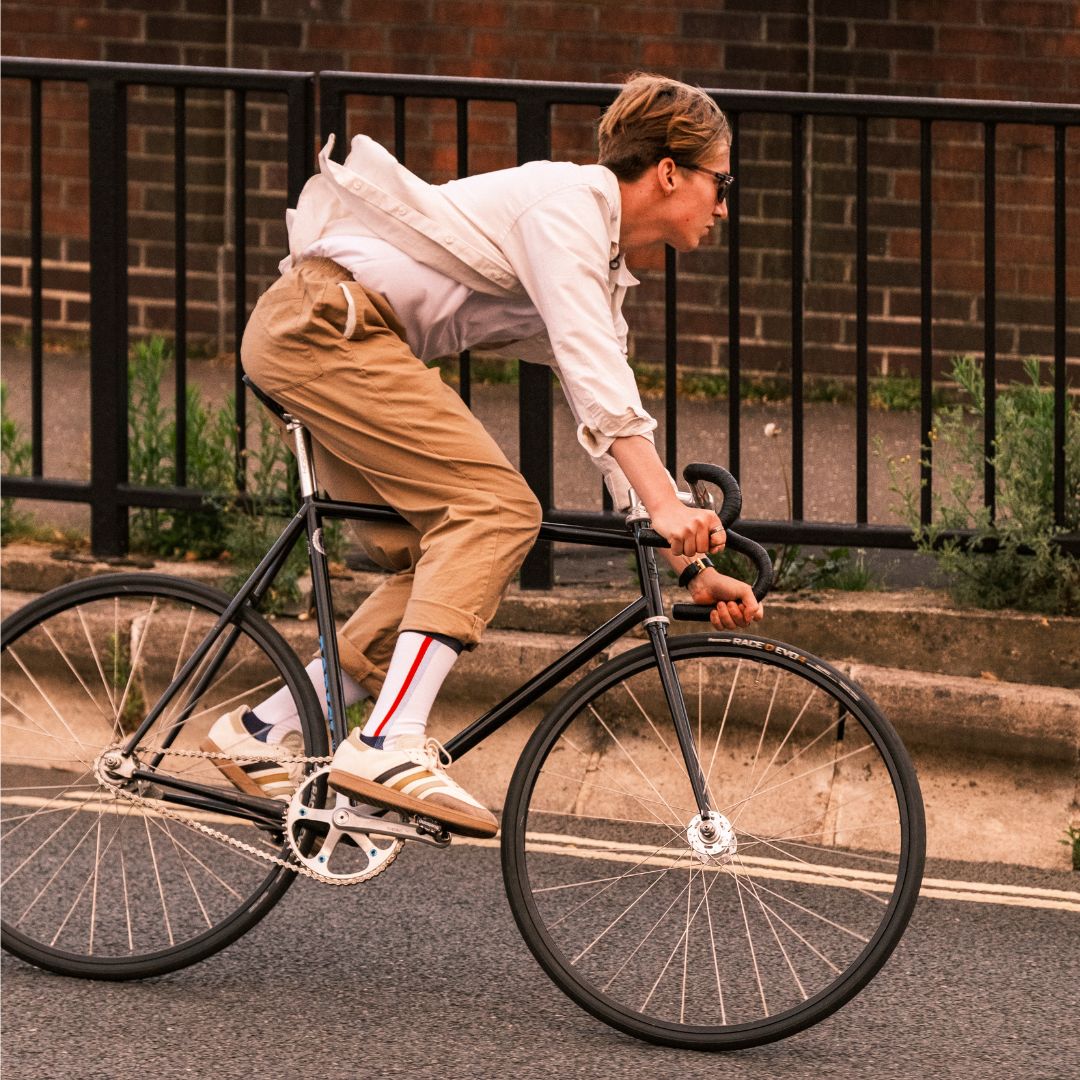 A person wearing a white shirt, khaki pants, sneakers, and striped socks rides a black bicycle on the street next to a metal fence and brick wall.