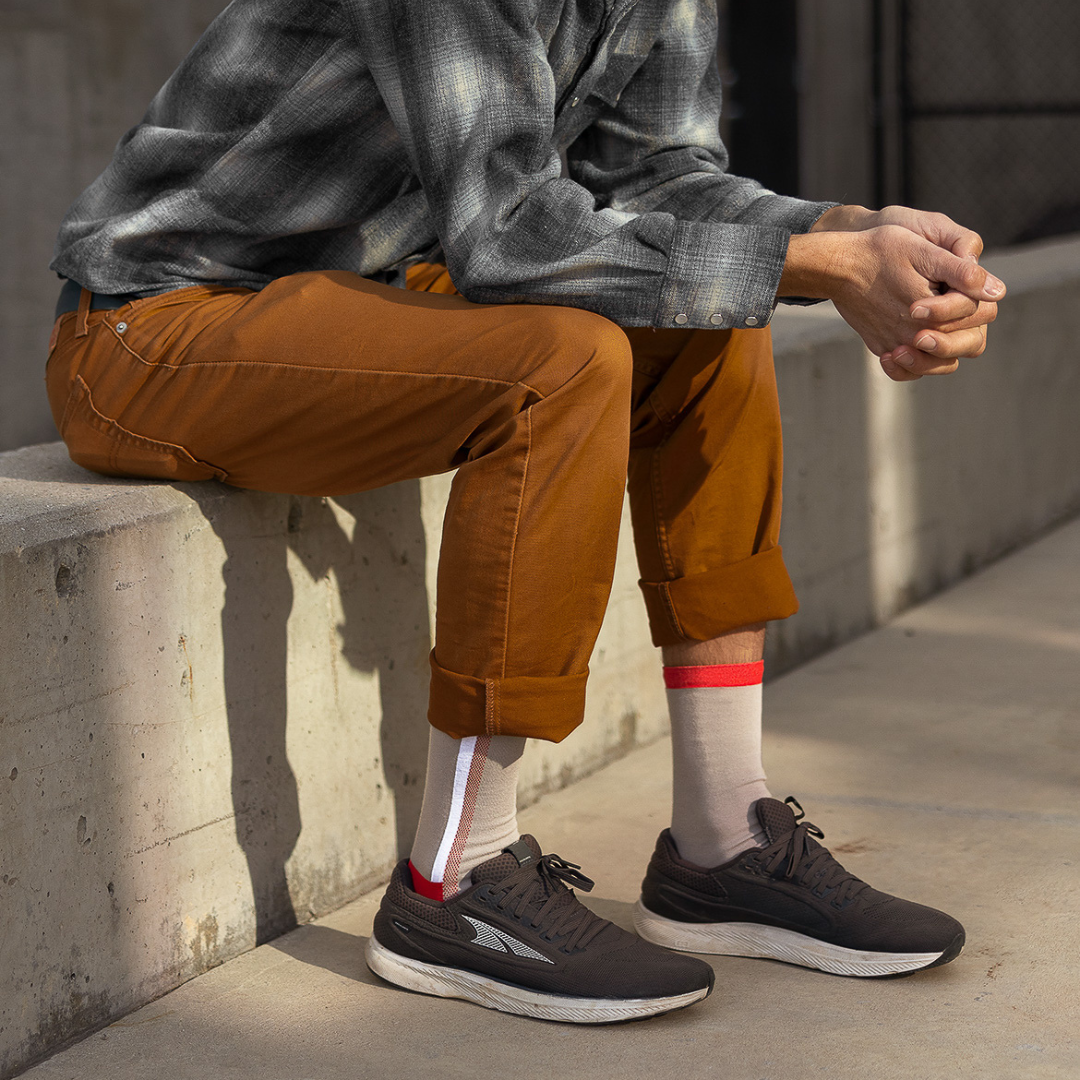 A person in a gray plaid shirt and rolled-up brown pants sits on a concrete bench, with their hands clasped. They wear white socks with red and gray stripes and black athletic shoes. The background shows a sunlit outdoor area.