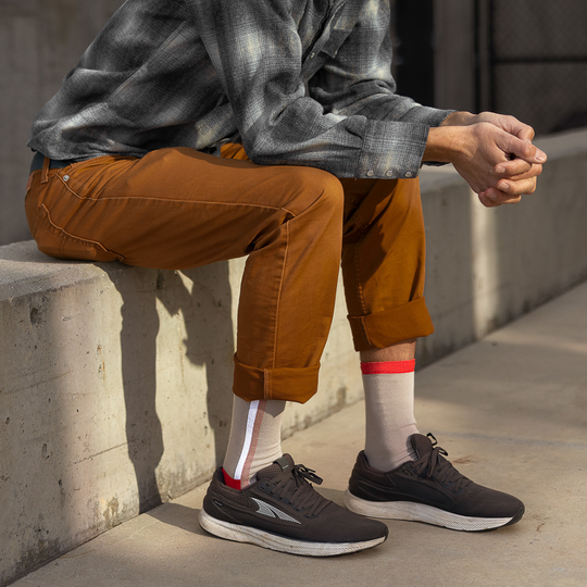A person in a gray plaid shirt and rolled-up brown pants sits on a concrete bench, with their hands clasped. They wear white socks with red and gray stripes and black athletic shoes. The background shows a sunlit outdoor area.
