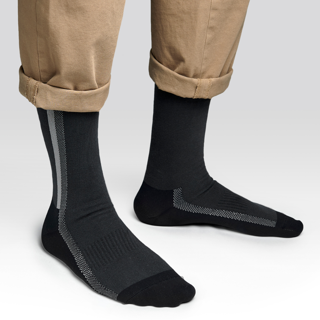 A person wearing tan pants with cuffs rolled up and black socks featuring white and gray stripe patterns along the sides and top, standing on a light gray background.