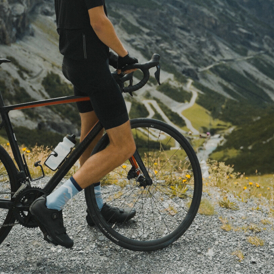 A cyclist in black gear stands beside a road bike on a mountainside, surrounded by wildflowers and rugged terrain, with winding roads and cliffs visible in the background.