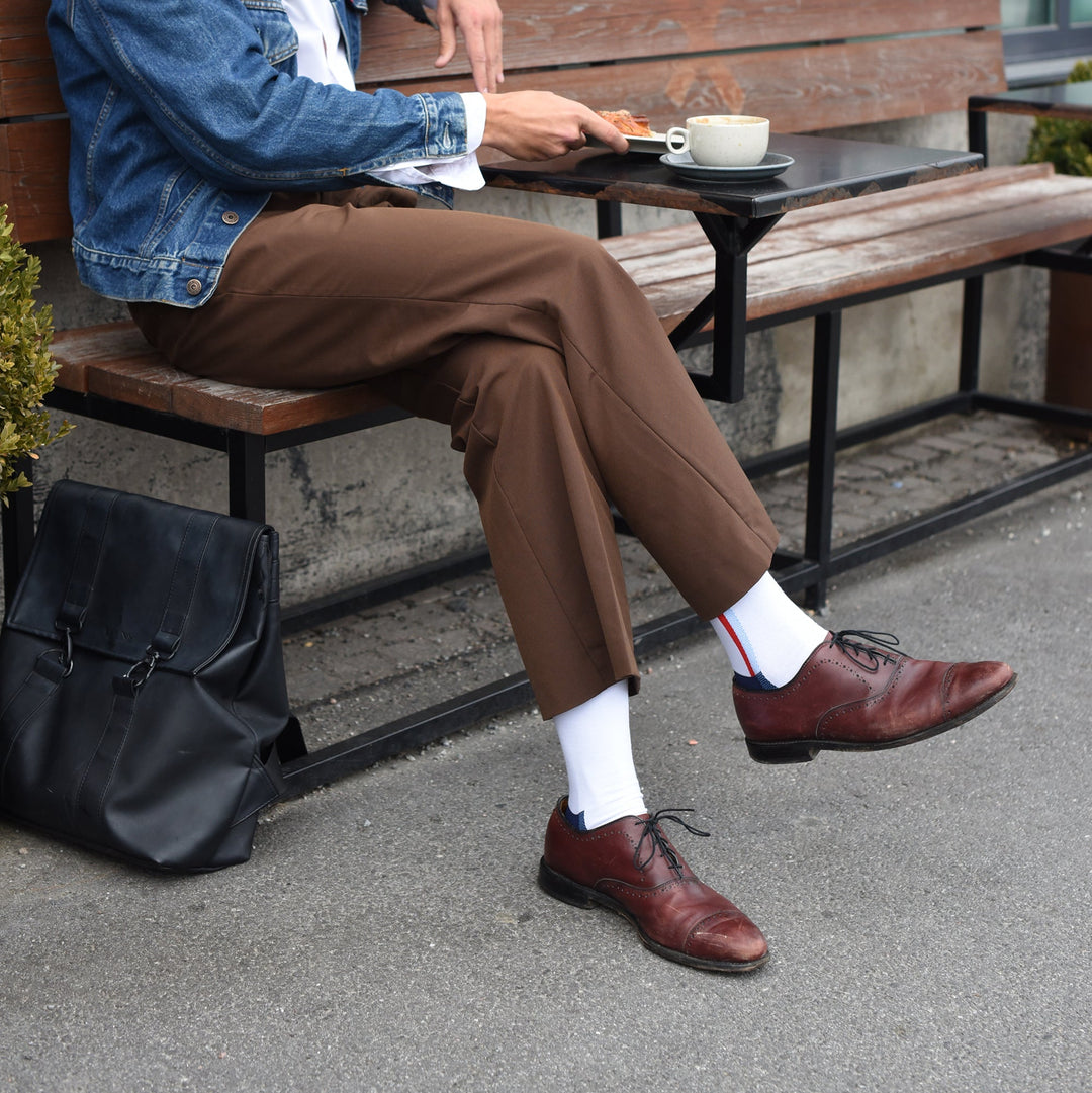 A person sits at an outdoor café table, wearing brown trousers, a denim jacket, white socks, and brown dress shoes. A black leather bag rests on the ground beside them, and there is a cup of coffee on the table.