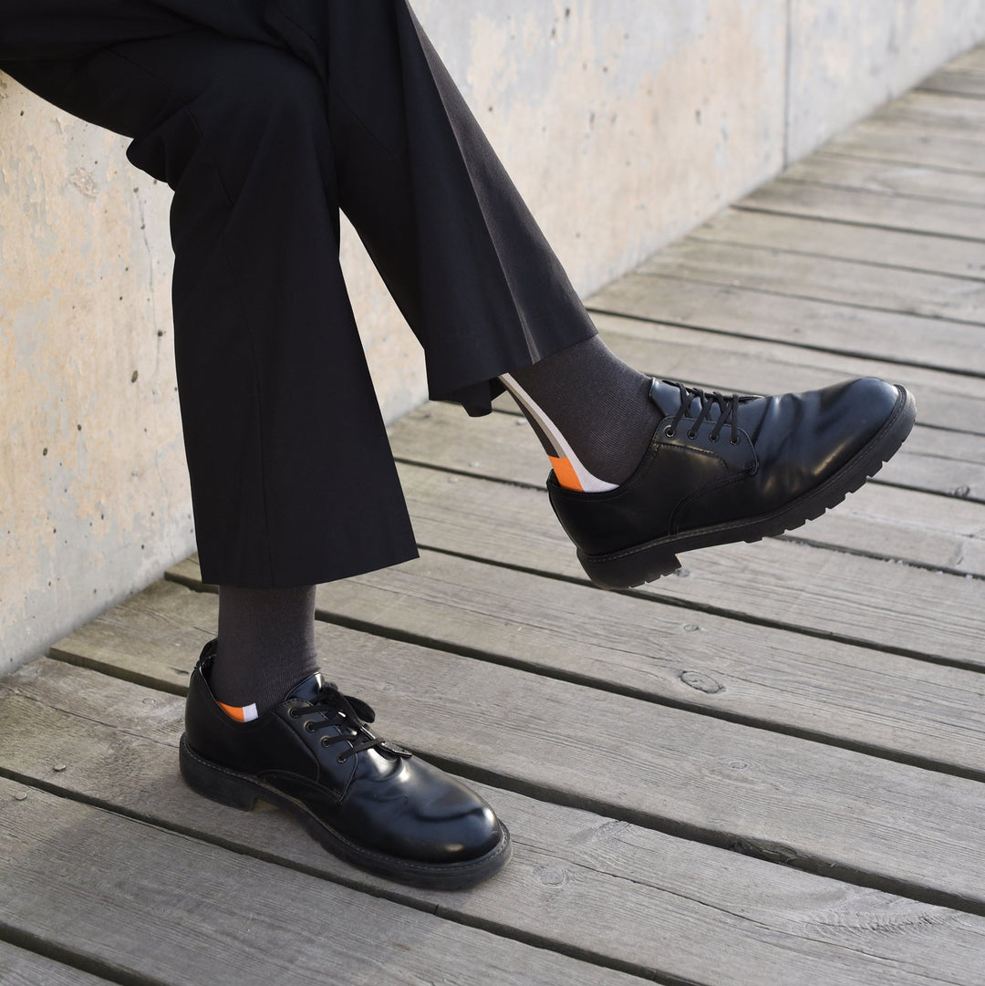 A person wearing black dress pants, dark socks with an orange and white stripe, and shiny black dress shoes sits on a wooden boardwalk with legs crossed.