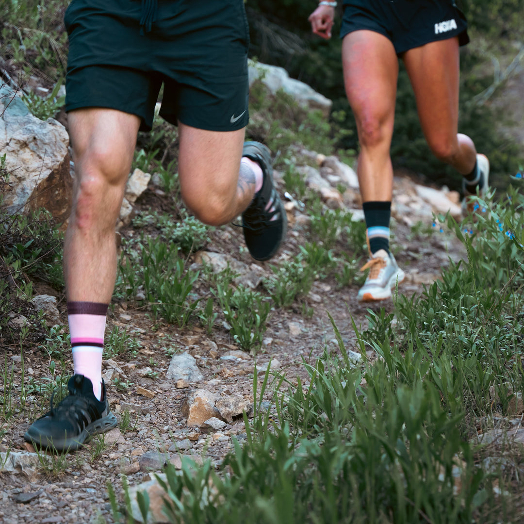 Two people trail running on a rocky, dirt path surrounded by greenery; only their legs are visible, each wearing shorts, socks, and running shoes.