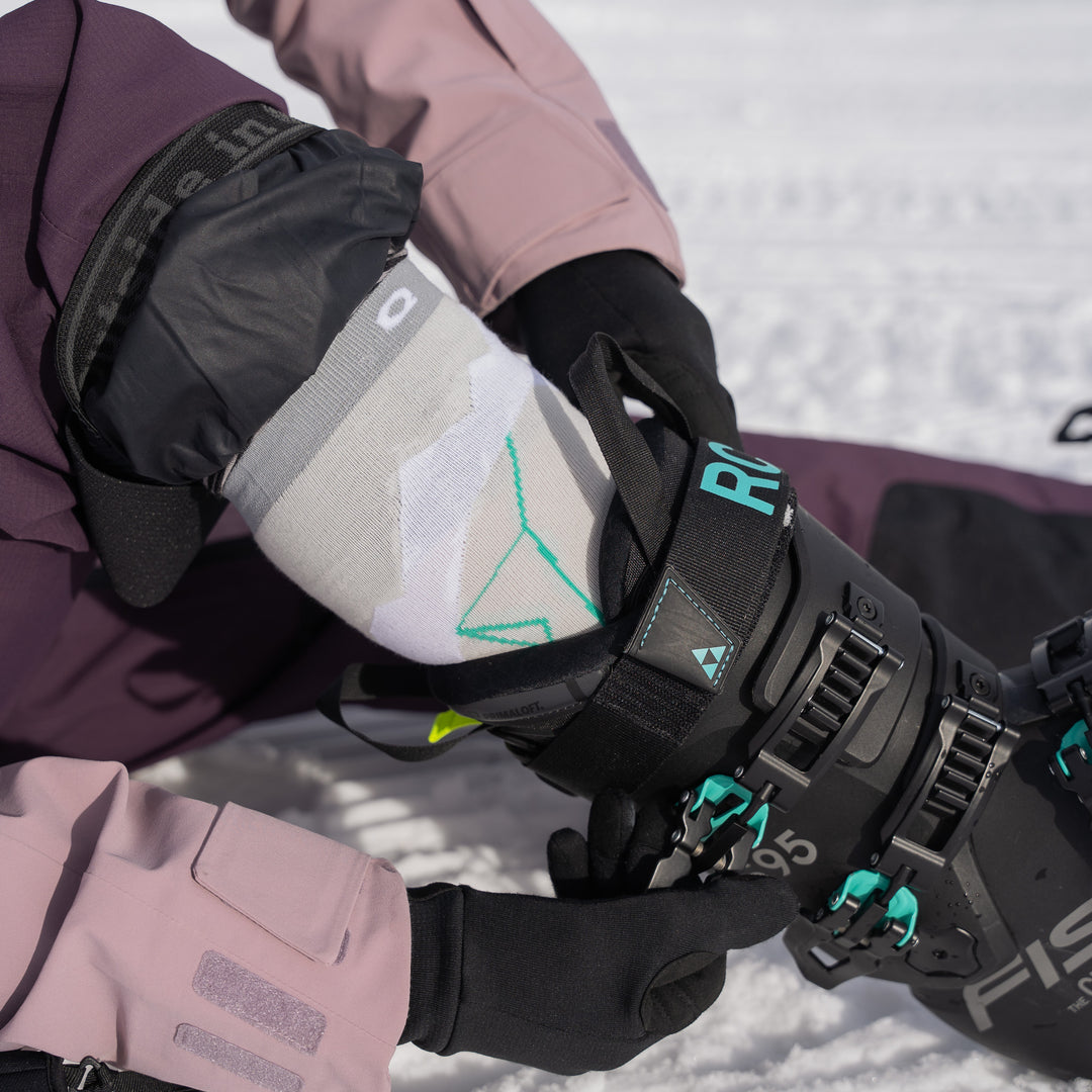 A person wearing a pink and black ski outfit adjusts the buckle of a black ski boot with teal accents while sitting on snow. The persons gloved hands and geometric-patterned sock are visible.