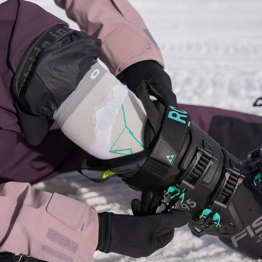 A person wearing a pink and black ski outfit adjusts the buckle of a black ski boot with teal accents while sitting on snow. The persons gloved hands and geometric-patterned sock are visible.