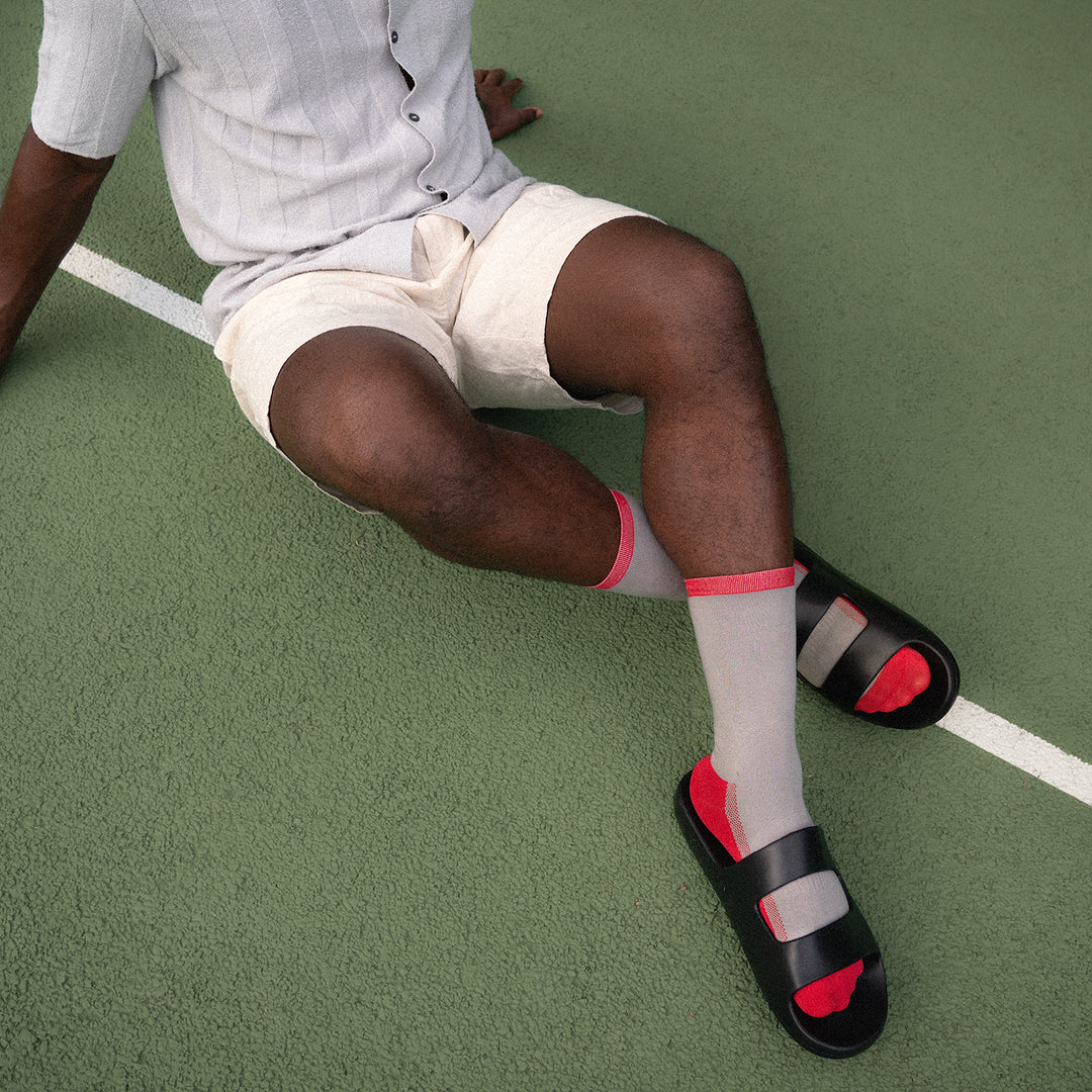 A person sits on a green tennis court, wearing a light gray shirt, beige shorts, red-and-gray socks, and black sandals. Only the lower half of their body is visible.
