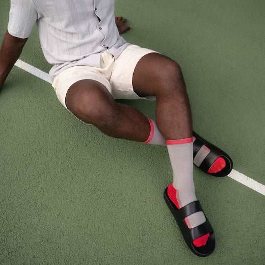 A person sits on a green tennis court, wearing a light gray shirt, beige shorts, red-and-gray socks, and black sandals. Only the lower half of their body is visible.