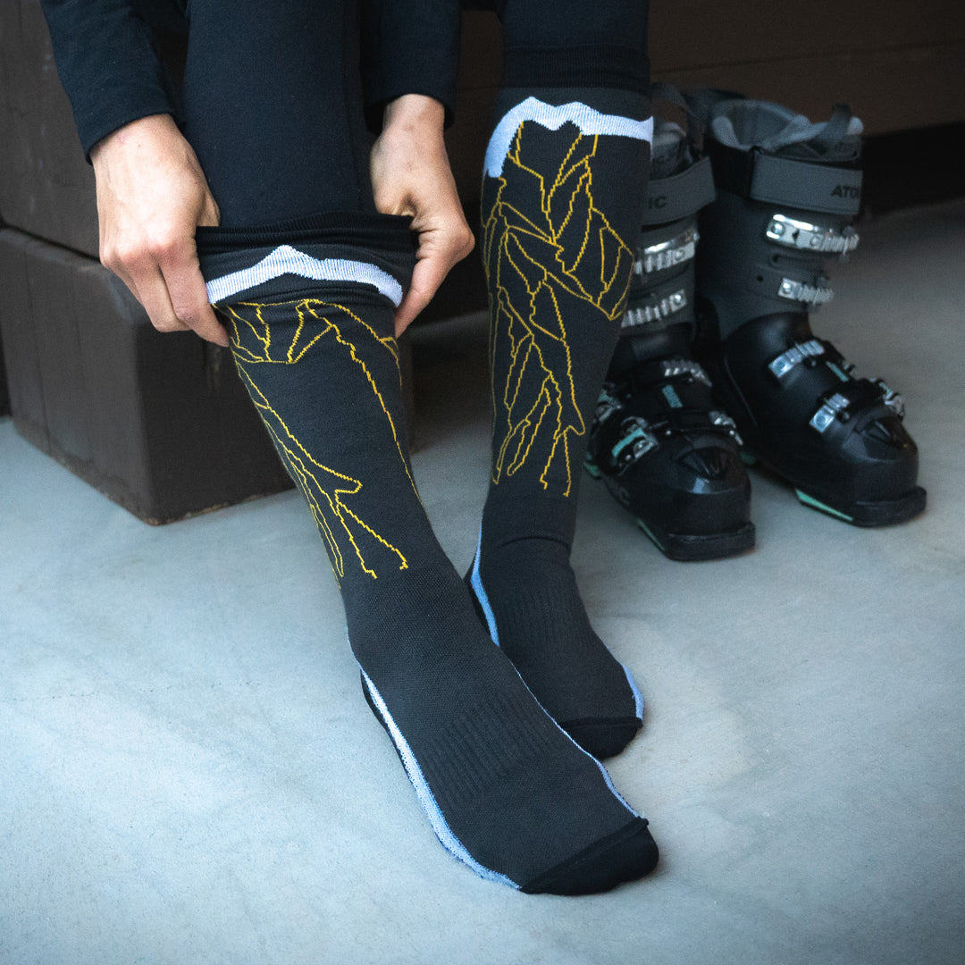A person pulls up black and yellow ski socks over their legs, with a pair of black ski boots placed on the floor nearby.
