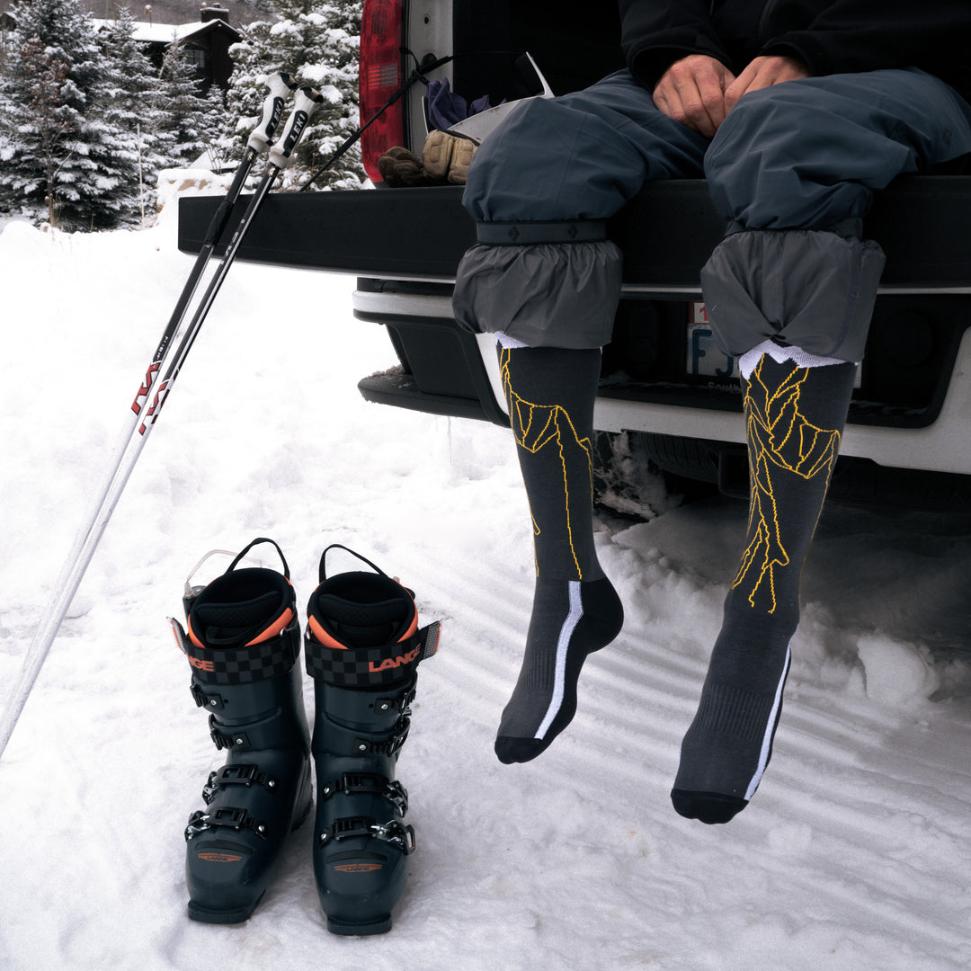 A person sits on the open tailgate of a truck in the snow, wearing long patterned ski socks and snow pants. Ski boots and poles are on the ground nearby, with snowy trees in the background.