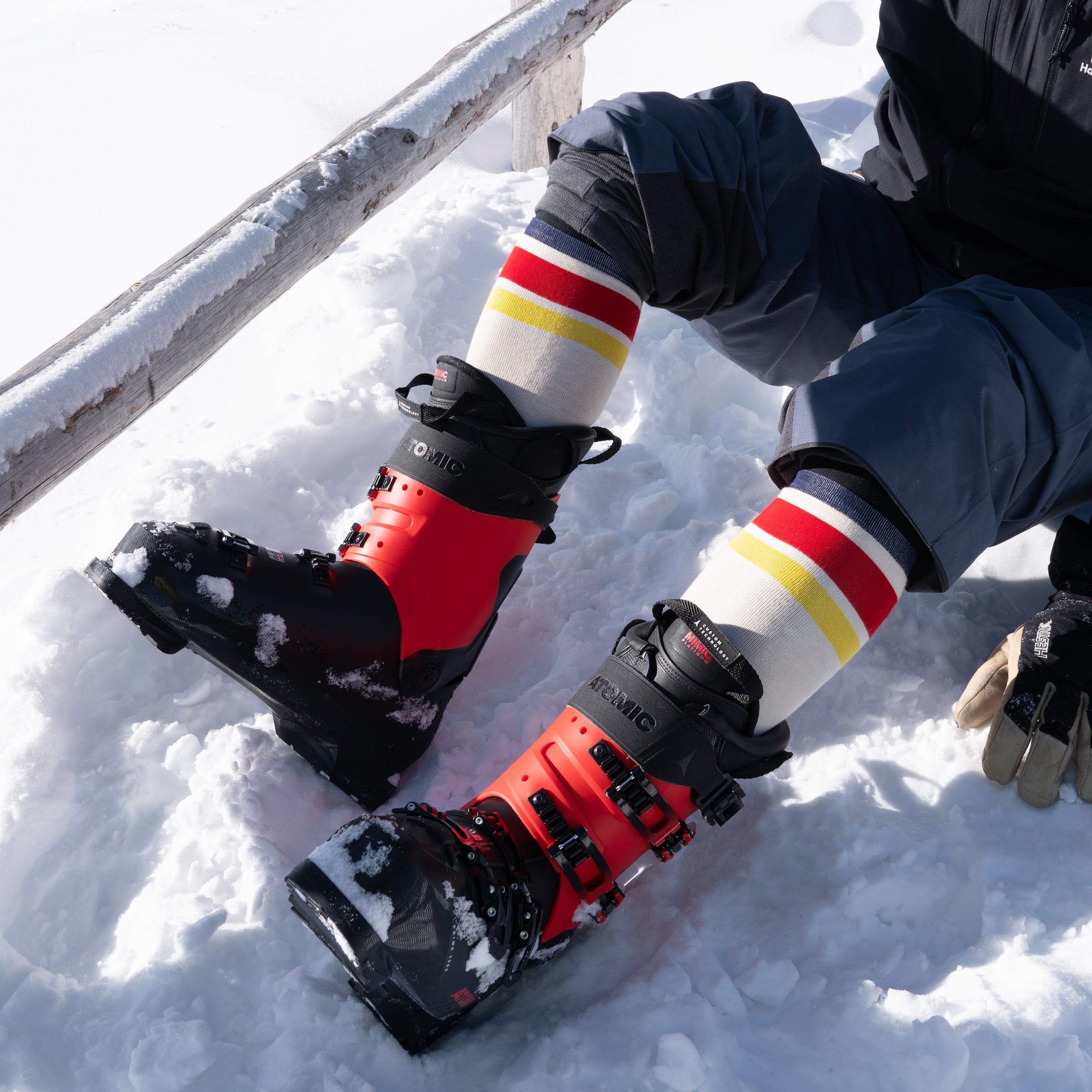 Person sitting in the snow wearing red ski boots and colorful striped socks, with dark snow pants rolled up. Snow-covered gloves rest nearby on the ground. Bright sunlight casts shadows on the snow.