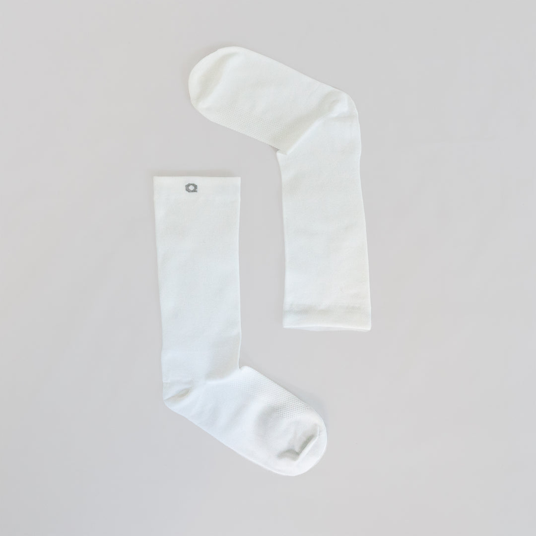 Two plain white socks laid flat on a light surface, positioned to form an angled shape resembling an upside-down letter L. One sock has a small gray circular emblem near the top.