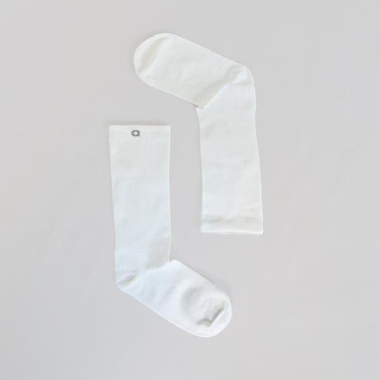 Two plain white socks laid flat on a light surface, positioned to form an angled shape resembling an upside-down letter L. One sock has a small gray circular emblem near the top.