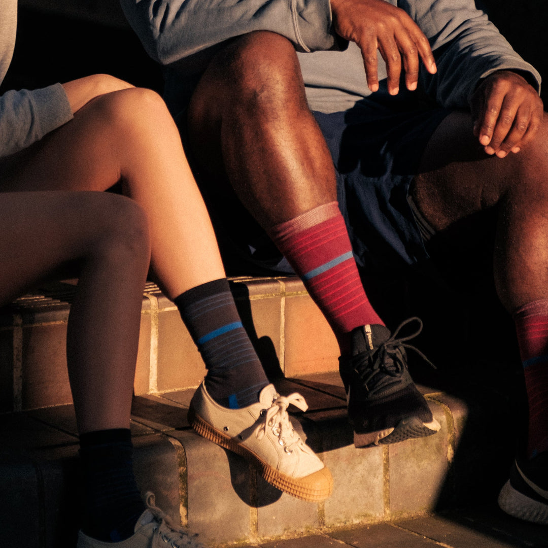 Two people sitting side by side on outdoor steps, wearing shorts, colorful mismatched socks, and sneakers. Their faces are not visible; only their legs and hands are shown in warm sunlight.