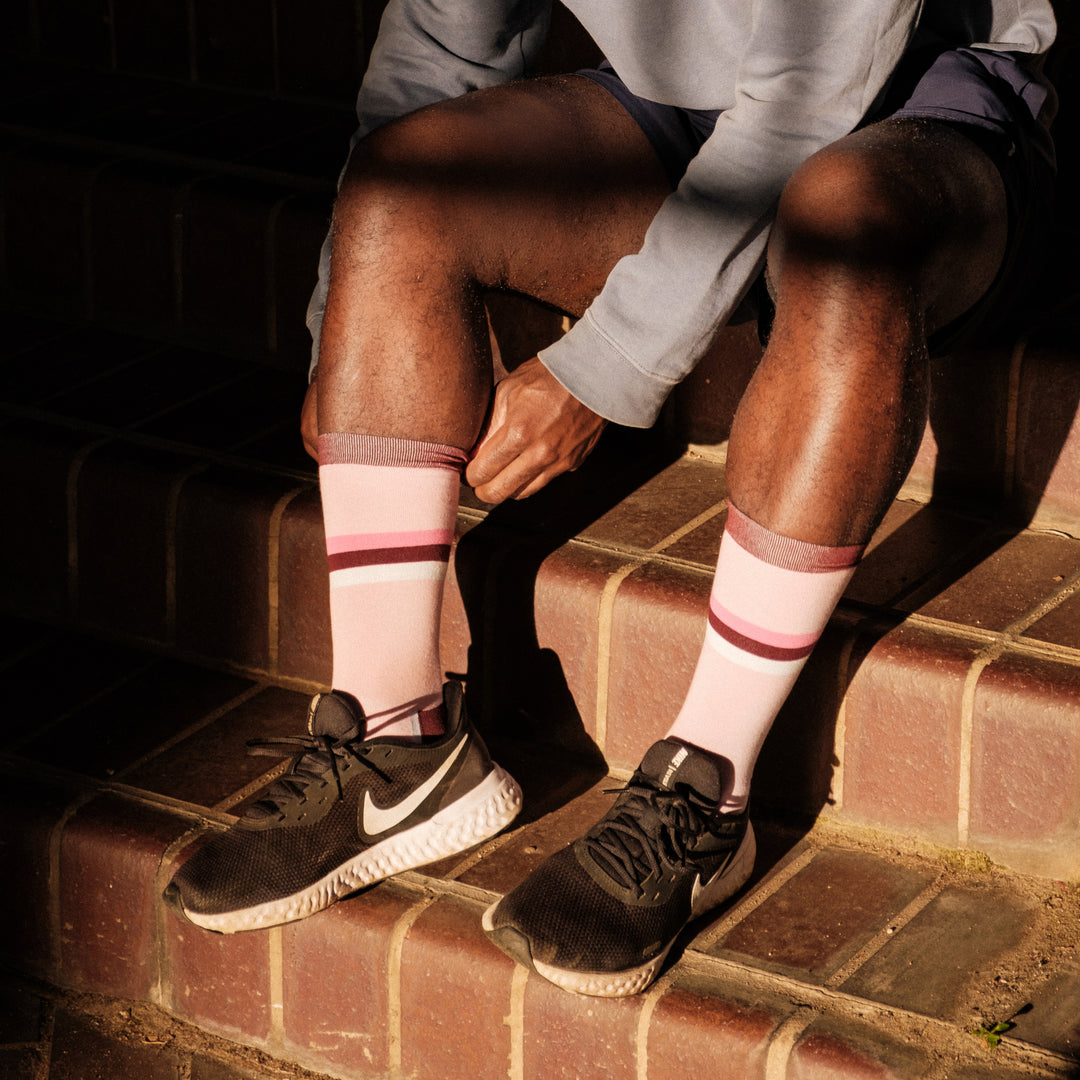 A person sitting on brick steps wears black Nike sneakers and pulls up pink socks with maroon stripes. Sunlight and shadows fall across their legs and hands, which are visible from mid-thigh down.