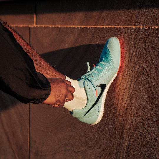 A person wearing black pants and white socks adjusts a light blue Nike running shoe with a red sole, standing against a textured brown stone surface in warm lighting.