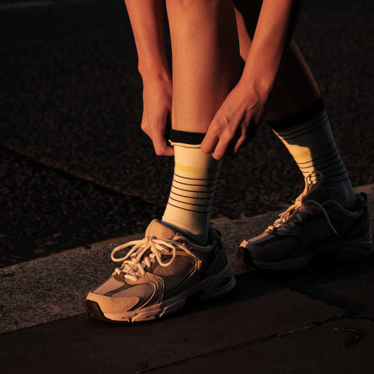 A person standing on a sidewalk adjusts their striped white sock, wearing gray and white sneakers. The scene is lit by warm, low sunlight.