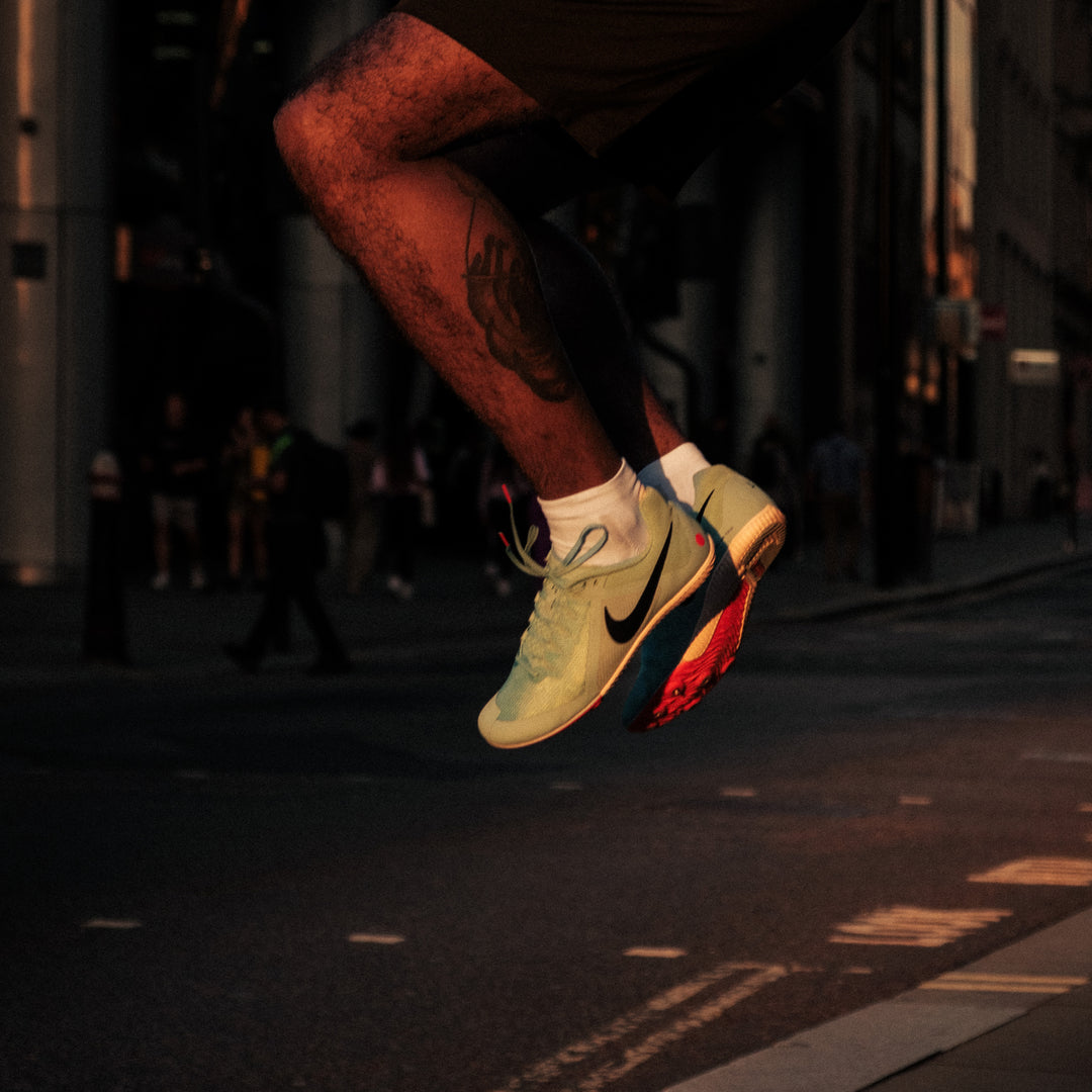 A person mid-jump over a crosswalk in an urban setting, wearing light green Nike sneakers with a red and blue sole; only their lower legs and feet are visible.