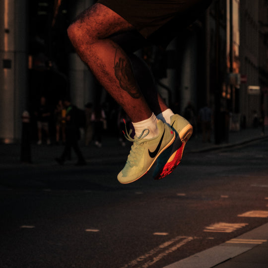 A person mid-jump over a crosswalk in an urban setting, wearing light green Nike sneakers with a red and blue sole; only their lower legs and feet are visible.