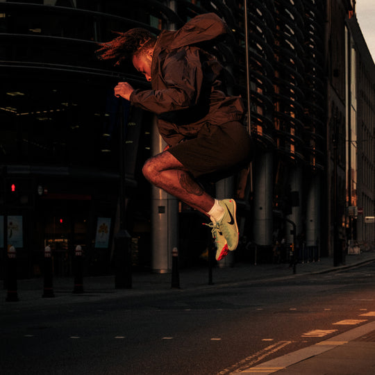 A person wearing a dark jacket, shorts, and light green sneakers is captured mid-air while jumping on an empty city street at dusk.