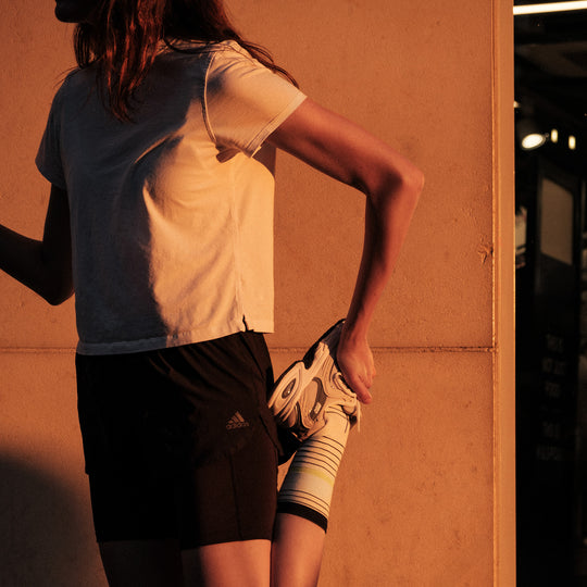 A person wearing a white t-shirt and black shorts stretches their leg by holding their foot behind them, standing against a wall in warm, golden sunlight.