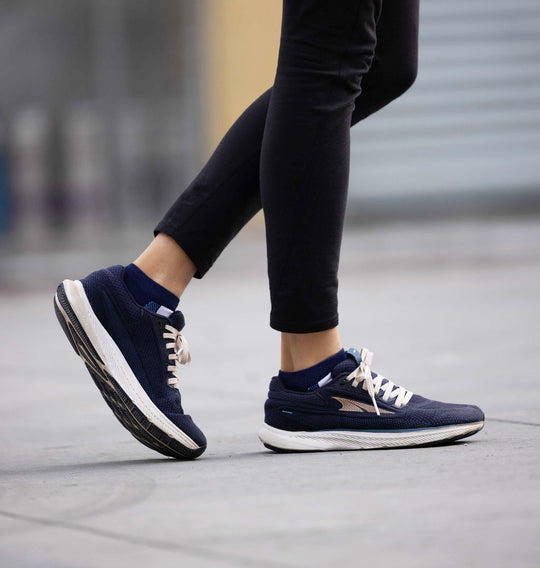 A person wearing navy blue running shoes with white soles and cream laces is walking on a sidewalk. Only their lower legs and feet are visible, dressed in black pants.