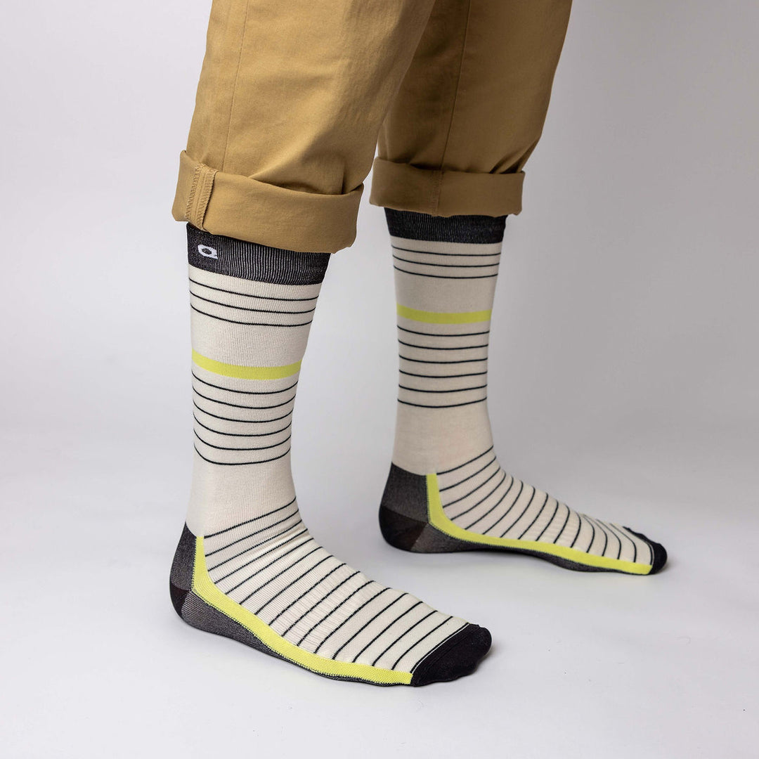 A person wearing tan pants rolled at the cuffs and cream-colored socks with black horizontal stripes, gray heels and toes, and yellow accents, standing on a plain white background.