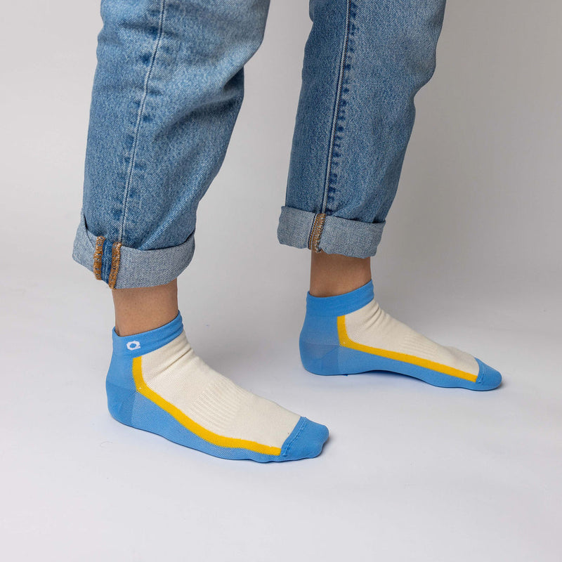 A person wearing rolled-up blue jeans and ankle socks with blue toes and heels, white tops, and yellow stripes, standing on a light-colored floor.