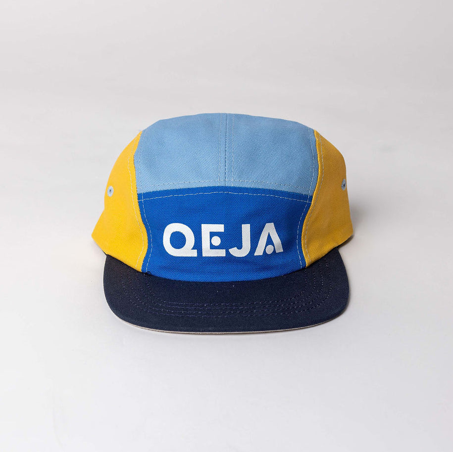 A five-panel cap with yellow and blue fabric panels, a dark blue brim, and the white text QEJA printed on the front panel. The cap is displayed against a plain, light background.