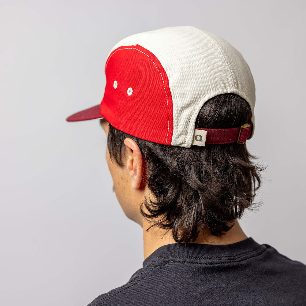 A person with dark, wavy hair wears a red and white baseball cap backward and a black shirt, facing away from the camera against a plain, light background.