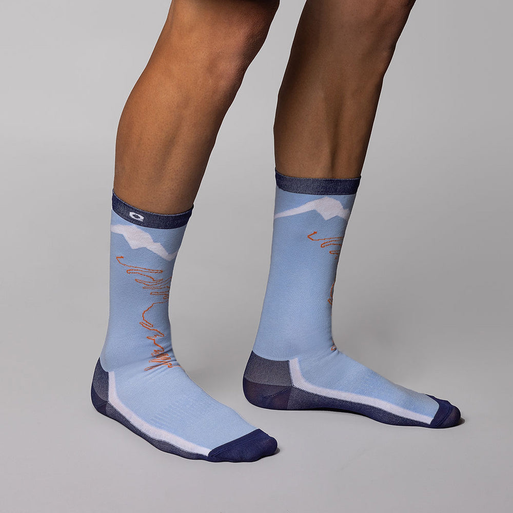 A person wears light blue crew socks with mountain and orange line designs, standing on a gray background.