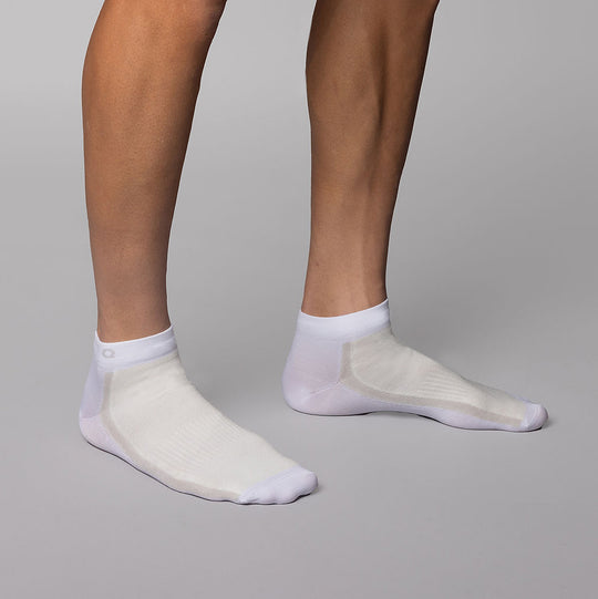 A person wearing white low-cut athletic socks stands on a light gray surface with feet slightly apart, showing only their lower legs and feet.