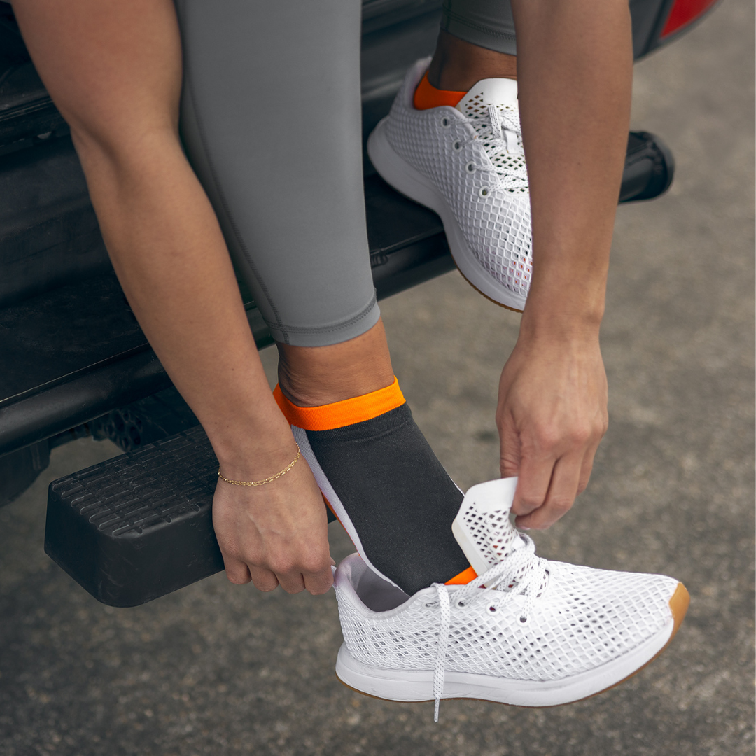 A person wearing gray leggings and black socks with orange and white stripes ties the laces of a white athletic shoe while sitting on the edge of a vehicle.