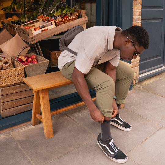 A man sitting on a wooden bench outside a produce shop adjusts his sock. He wears glasses, a light polo shirt, olive green pants, black sneakers, and a gray crossbody bag. Wooden crates with vegetables are in the background.