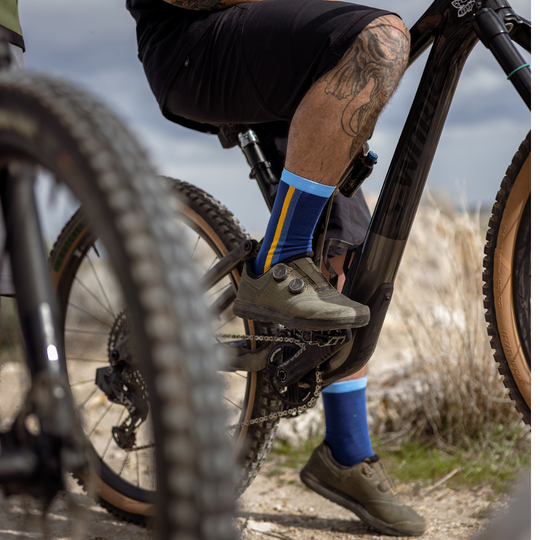A close-up of a cyclist’s tattooed legs on a mountain bike, wearing blue and yellow socks and dark shoes, with another bike tire visible in the foreground on a rocky outdoor trail.
