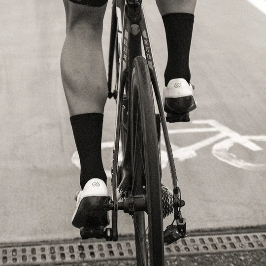 Black and white photo taken from behind of a cyclist riding on a road, showing their legs, socks, cycling shoes, bike frame, and a bike lane symbol painted on the pavement.
