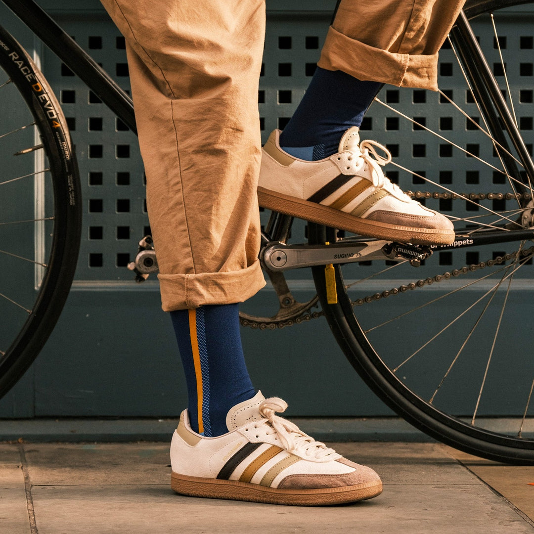 A person wearing khaki pants, blue socks with a yellow stripe, and white sneakers with brown and gold stripes stands next to a black bicycle. The pants are rolled up and the bike has a visible rear wheel and pedal.